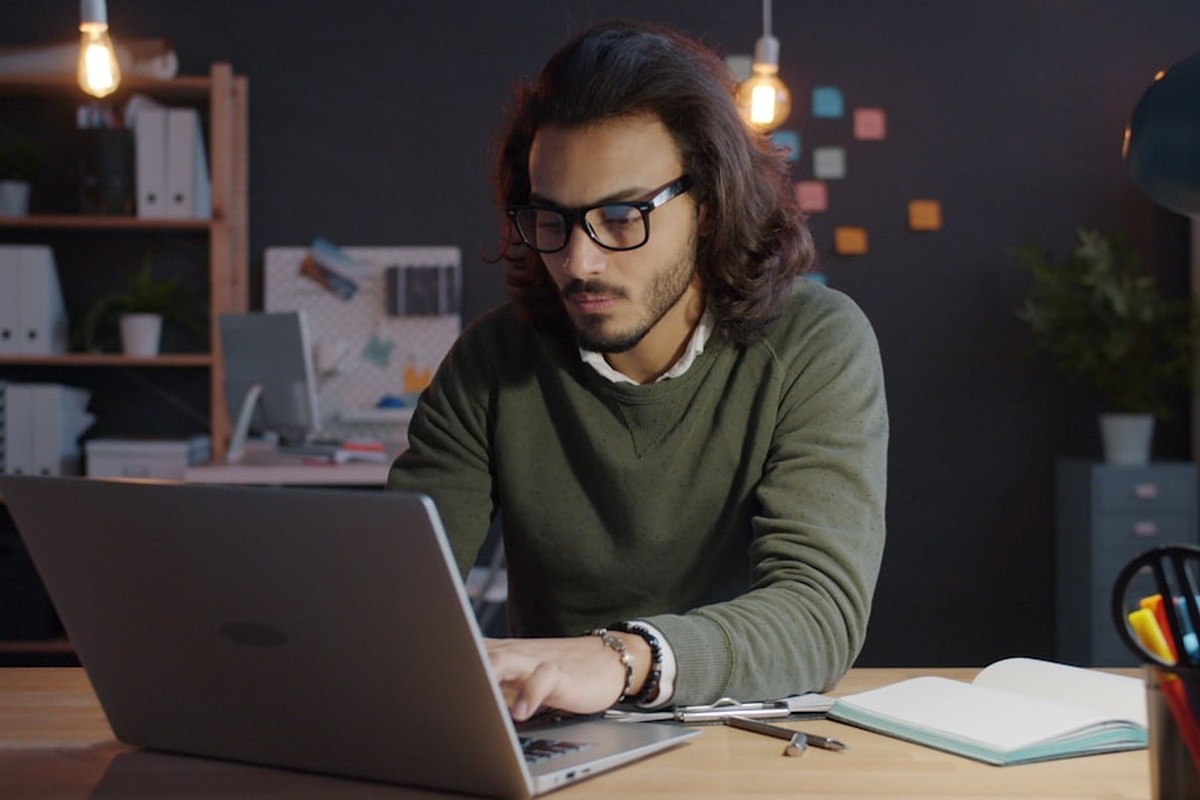 man standing and facing whiteboard with writings near woman pointing at it and facing man sitting beside table with laptop indoors