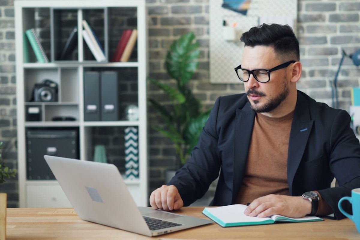 man standing and facing whiteboard with writings near woman pointing at it and facing man sitting beside table with laptop indoors